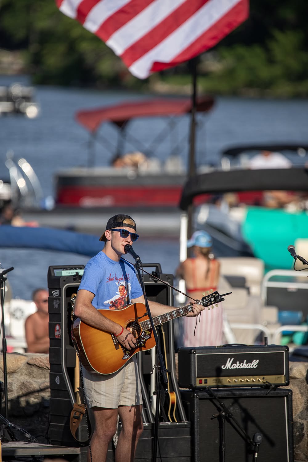 Ryan North performing acoustic guitar at an outdoor lakeside event with an American flag in the background